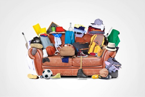 A brown fabric armchair fully covered with a variety of discarded items, including sports gear such as a soccer ball and a basketball, clothing items like shirts, trousers, and hats, as well as miscellaneous objects like a yellow toolbox, a green backpack, a red umbrella, and a white waste bin. The objects are jumbled together, spilling over the arms and seat of the chair, with some items resting on the floor in front of it. The background is plain white, emphasizing the cluttered collection of rubbish piled on and around the armchair, illustrating clutter disposal or rubbish removal in a domestic or commercial setting near Hanwell, London, relevant to services provided by Commercial Waste Hanwell.