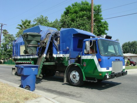 A large commercial rubbish collection truck parked on a suburban street, featuring a predominantly blue and green body with a metallic grey front cabin. The truck has a hydraulic lifting mechanism on the side, used for loading waste into the rear compartment, which is designed for carrying large quantities of refuse. The rear section of the vehicle contains a large, green, open-top compactor area with visible metal sides, and a green waste bin is positioned at the front of the truck, ready for collection. The street scene includes a concrete sidewalk and a grassy verge, with trees and utility poles in the background, indicating a residential or light-commercial area. Bright daylight illuminates the scene, highlighting the clean, industrial finish of the vehicle and the natural surroundings. The image aligns with rubbish removal services in the Hanwell area, demonstrating the type of equipment used by companies like Commercial Waste Hanwell for waste collection and disposal within the locality.