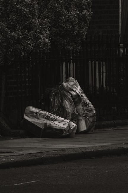 Two large black trash bags filled with waste are situated on a paved sidewalk in front of a black metal fence, which appears to have vertical bars and is partially obscured by dense foliage from a nea