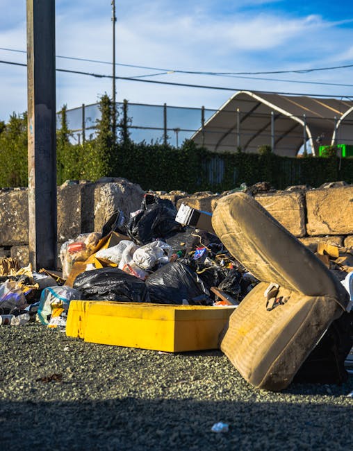Two large black trash bags filled with waste are situated on a paved sidewalk in front of a black metal fence, which appears to have vertical bars and is partially obscured by dense foliage from a nea