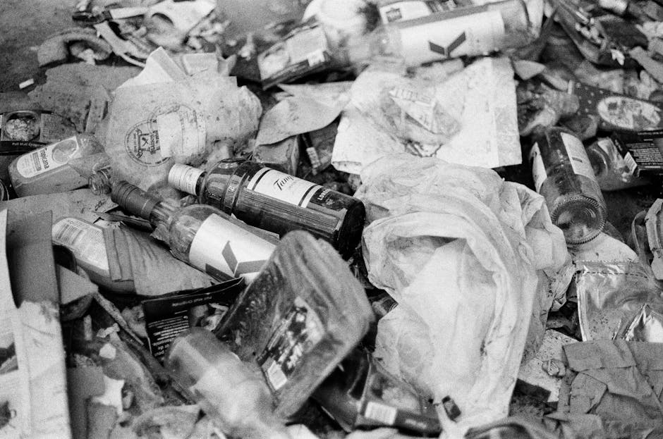 A small blue flatbed truck parked on the side of a street, loaded with a large quantity of mixed waste materials including flattened cardboard boxes, plastic bags, and various other discarded items. T
