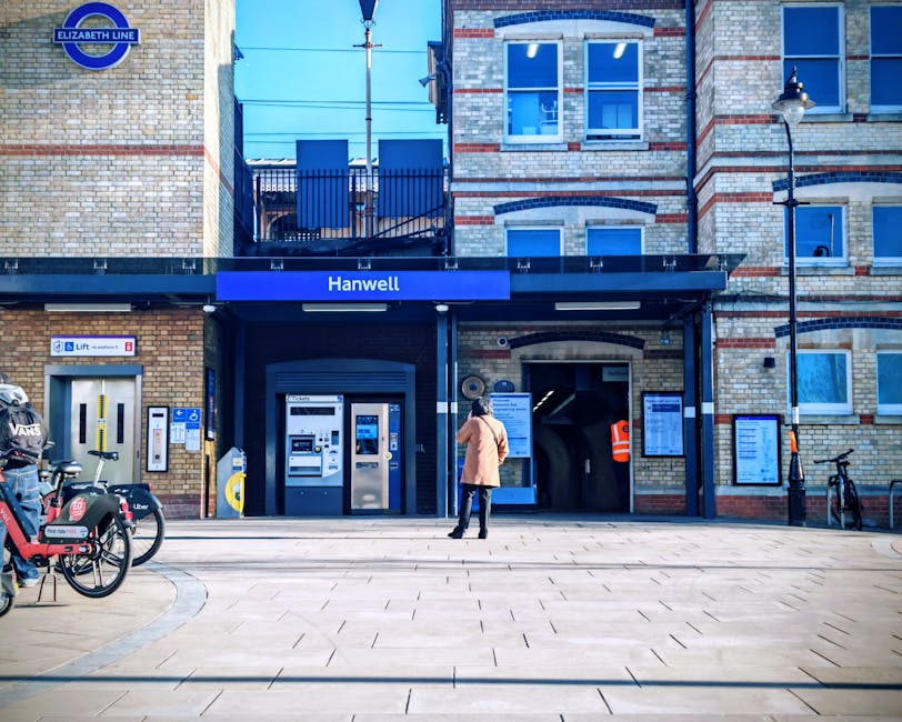 A street-level view of Hanwell railway station entrance showing a brick building with a blue sign labeled 'Hanwell' above the main doorway, which features ticket machines and a small entrance to the p