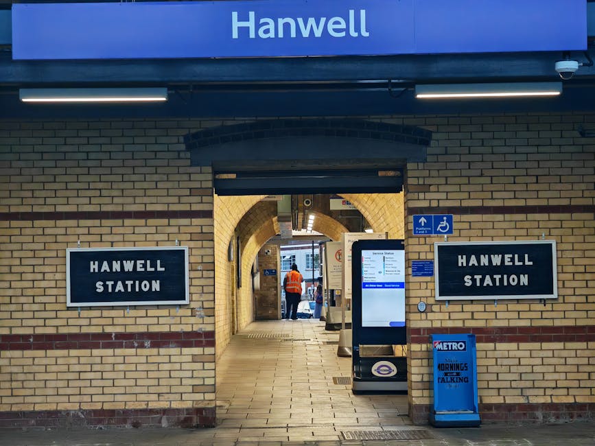 A street-level view of Hanwell railway station entrance showing a brick building with a blue sign labeled 'Hanwell' above the main doorway, which features ticket machines and a small entrance to the p